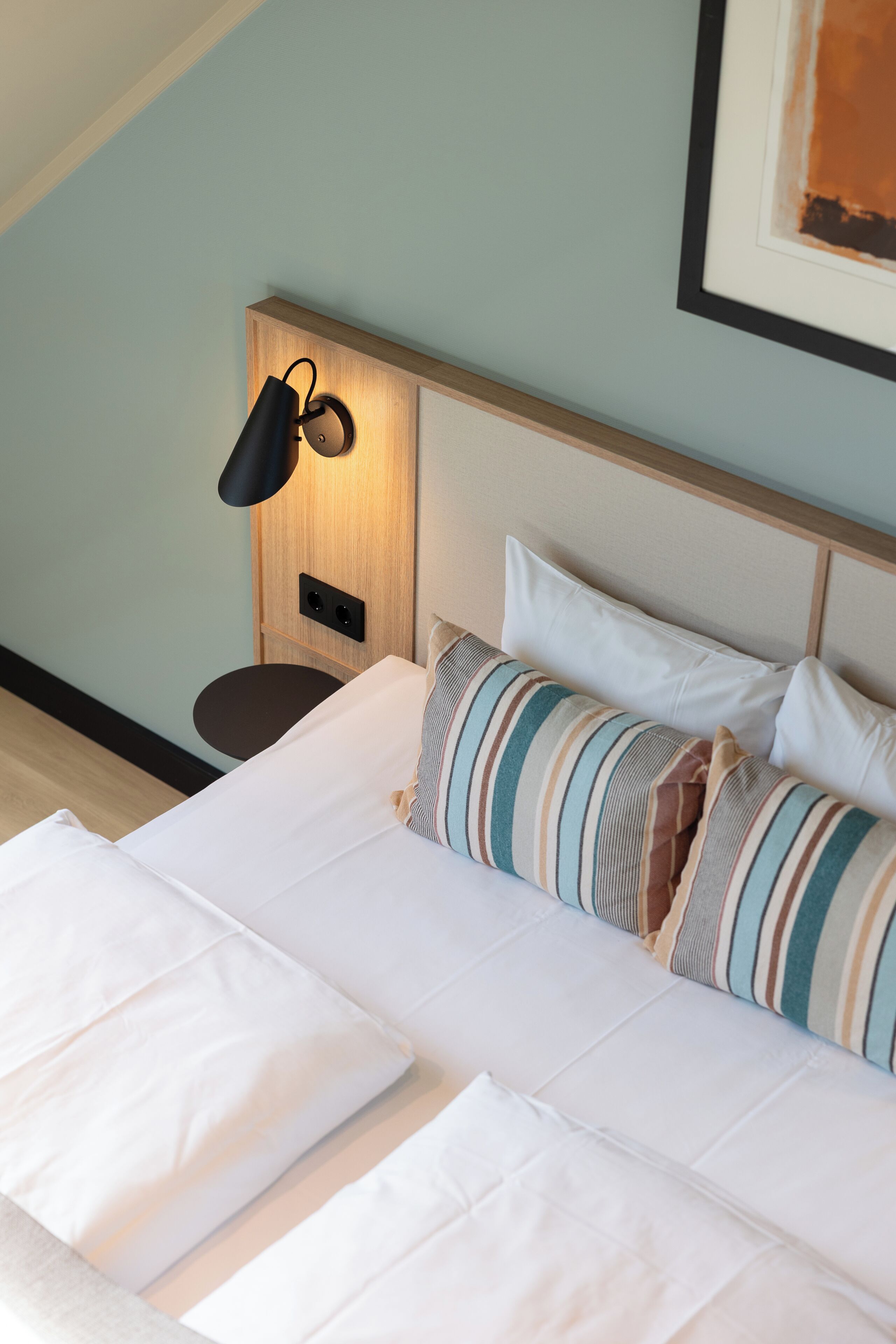 Una cama bien hecha con sábanas blancas y almohadas a rayas, una lámpara de noche y un cuadro en la pared.