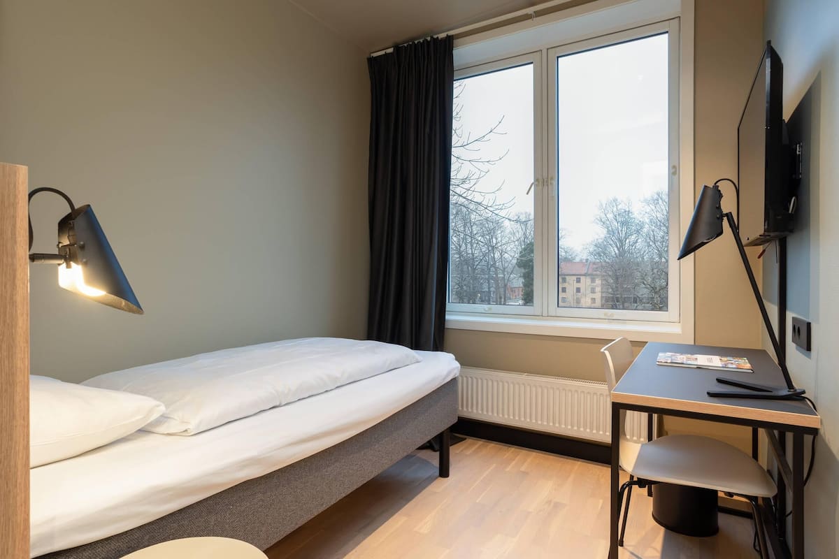 Ein Schlafzimmer mit einem Einzelbett, einem Schreibtisch, einem Stuhl, einem Fernseher und einem Fenster mit Vorhängen.