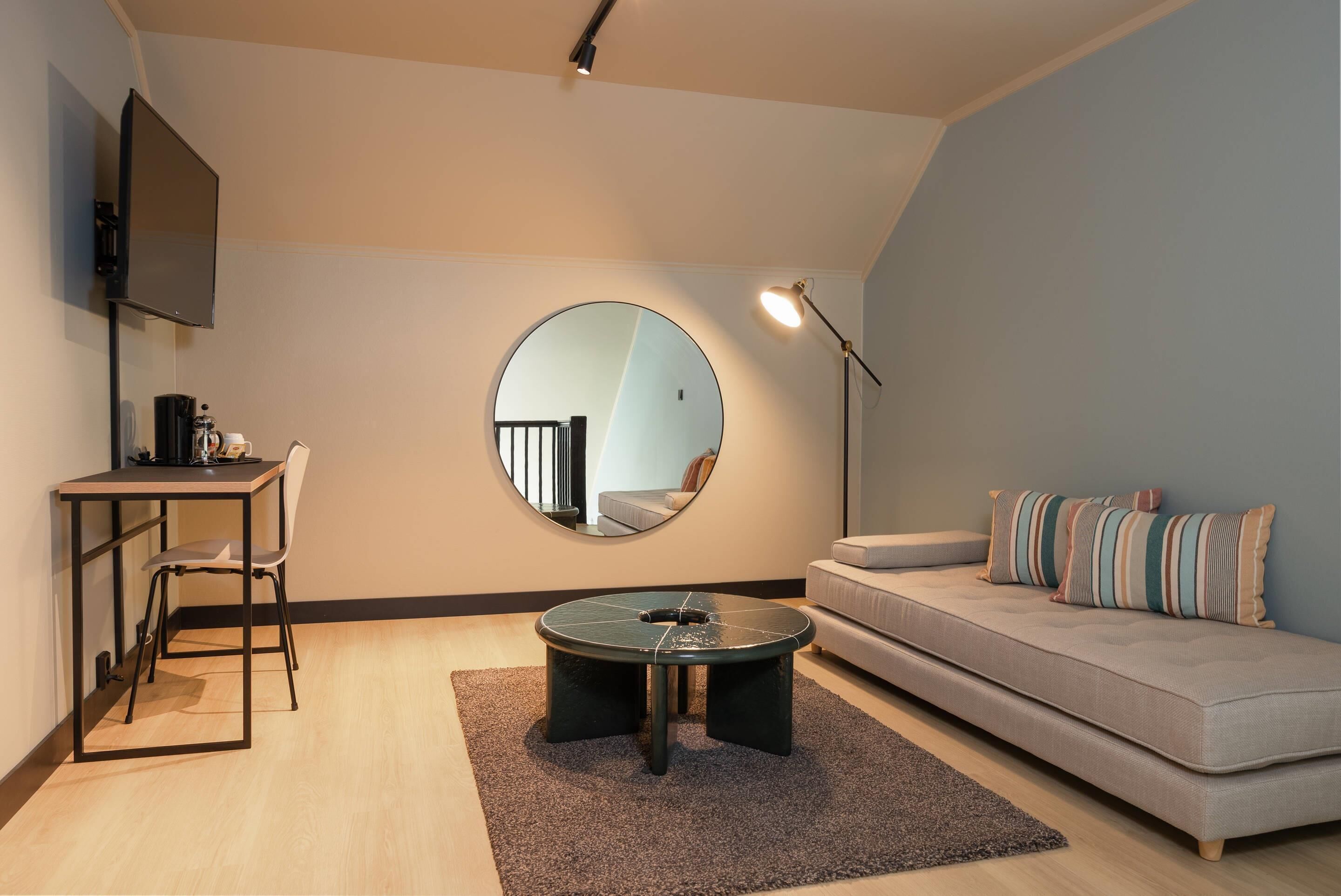 A modern living room with a round mirror, a sofa, a coffee table, a desk with a chair, and a TV.