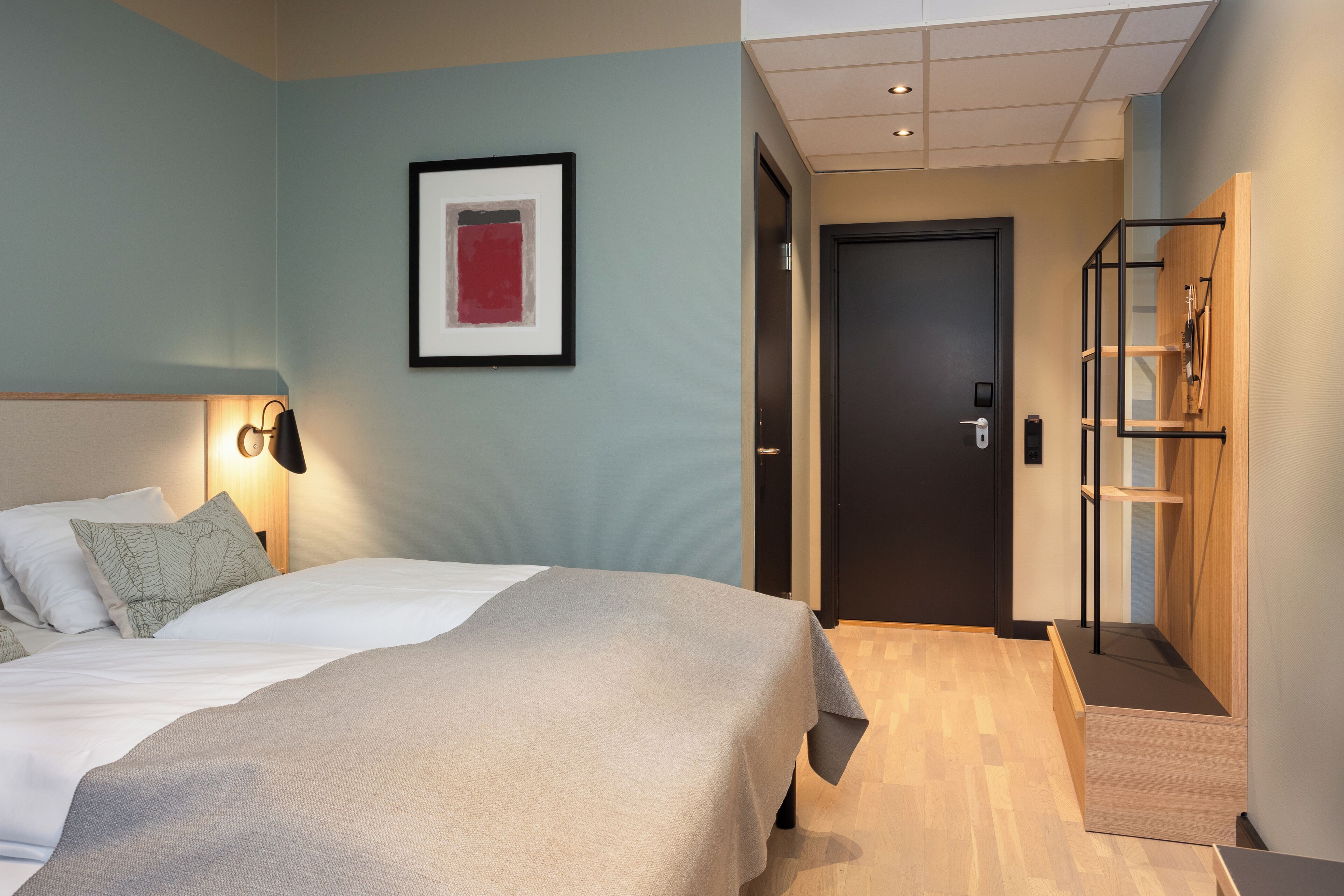 Moderni hotellihuone, jossa on sänky, yöpöytälamppu, kehystetty taideteos ja puinen vaatekaappi.
