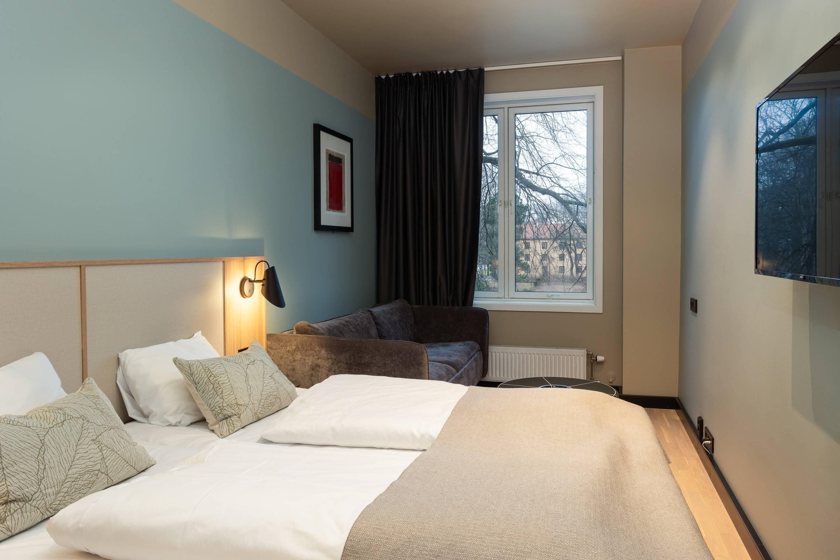 A hotel room with a bed, a sofa, a window with curtains, a wall-mounted TV, and a framed picture.