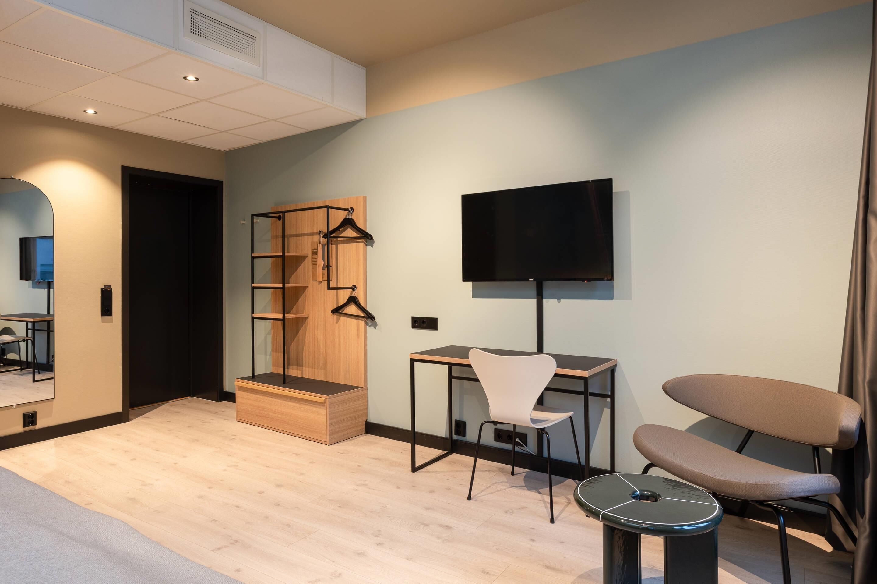 A modern hotel room with a wooden desk, a chair, a TV mounted on the wall, and a small seating area with a cushioned bench.