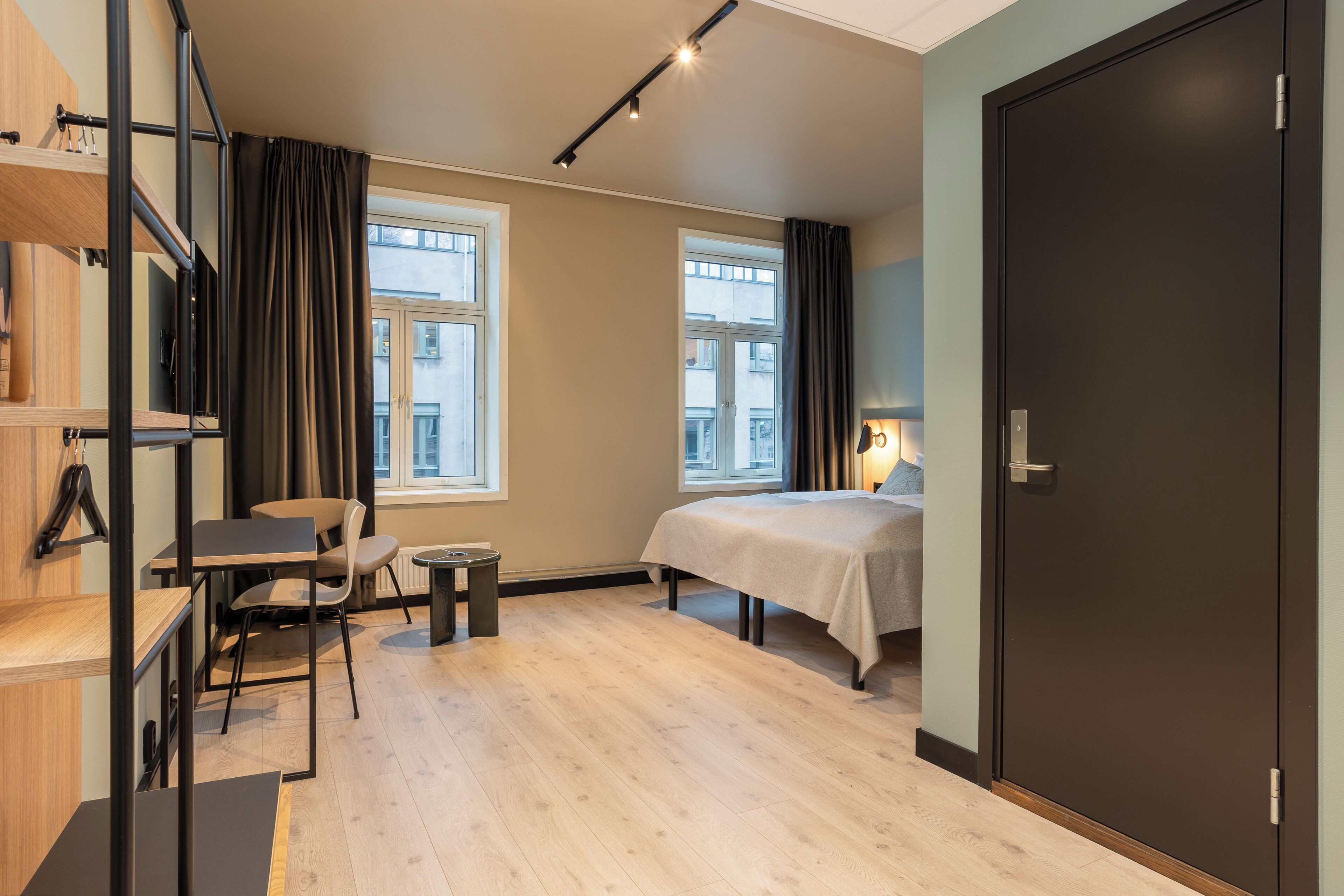 A modern hotel room with a bed, a desk, and a chair. There are two windows with curtains, a door, and a shelf with items.