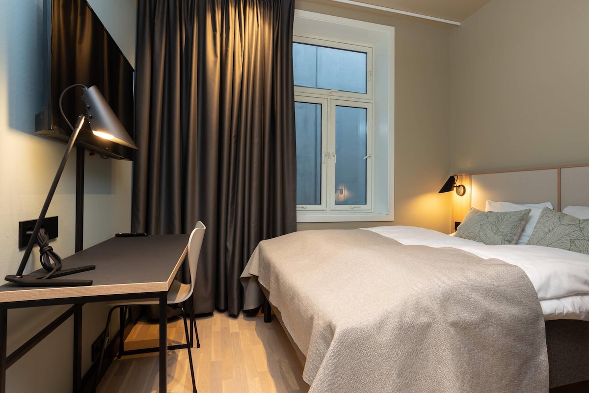 Ein Hotelzimmer mit einem Bett, einem Schreibtisch mit Lampe, einem Stuhl, einem Fenster mit Vorhängen und einem Nachttisch mit Lampe.