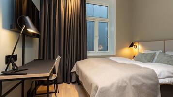 A hotel room with a bed, a desk with a lamp, a chair, a window with curtains, and a bedside table with a lamp.