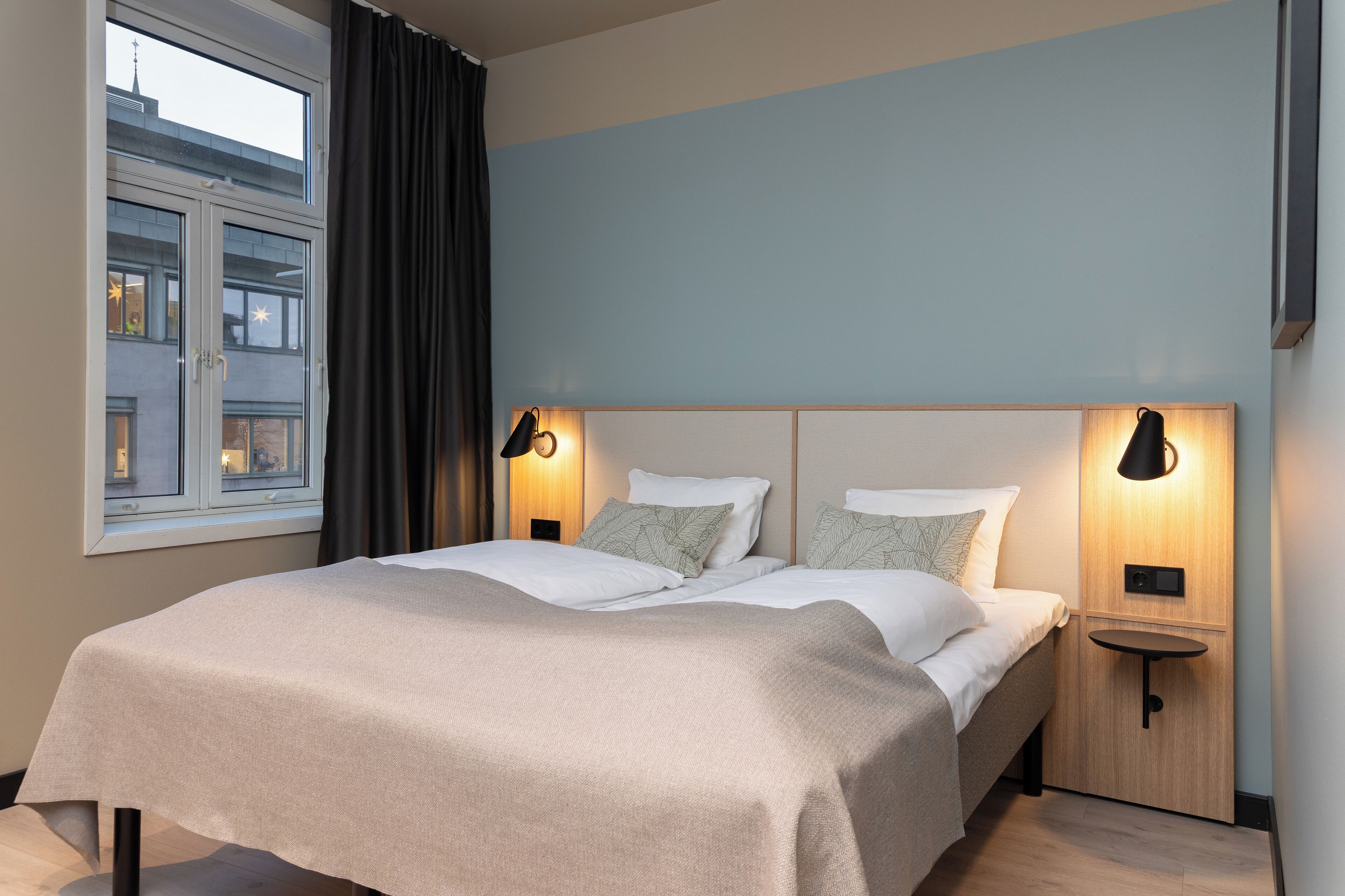 A modern bedroom with a large bed, two bedside tables with lamps, a window with curtains, and a view of buildings outside.