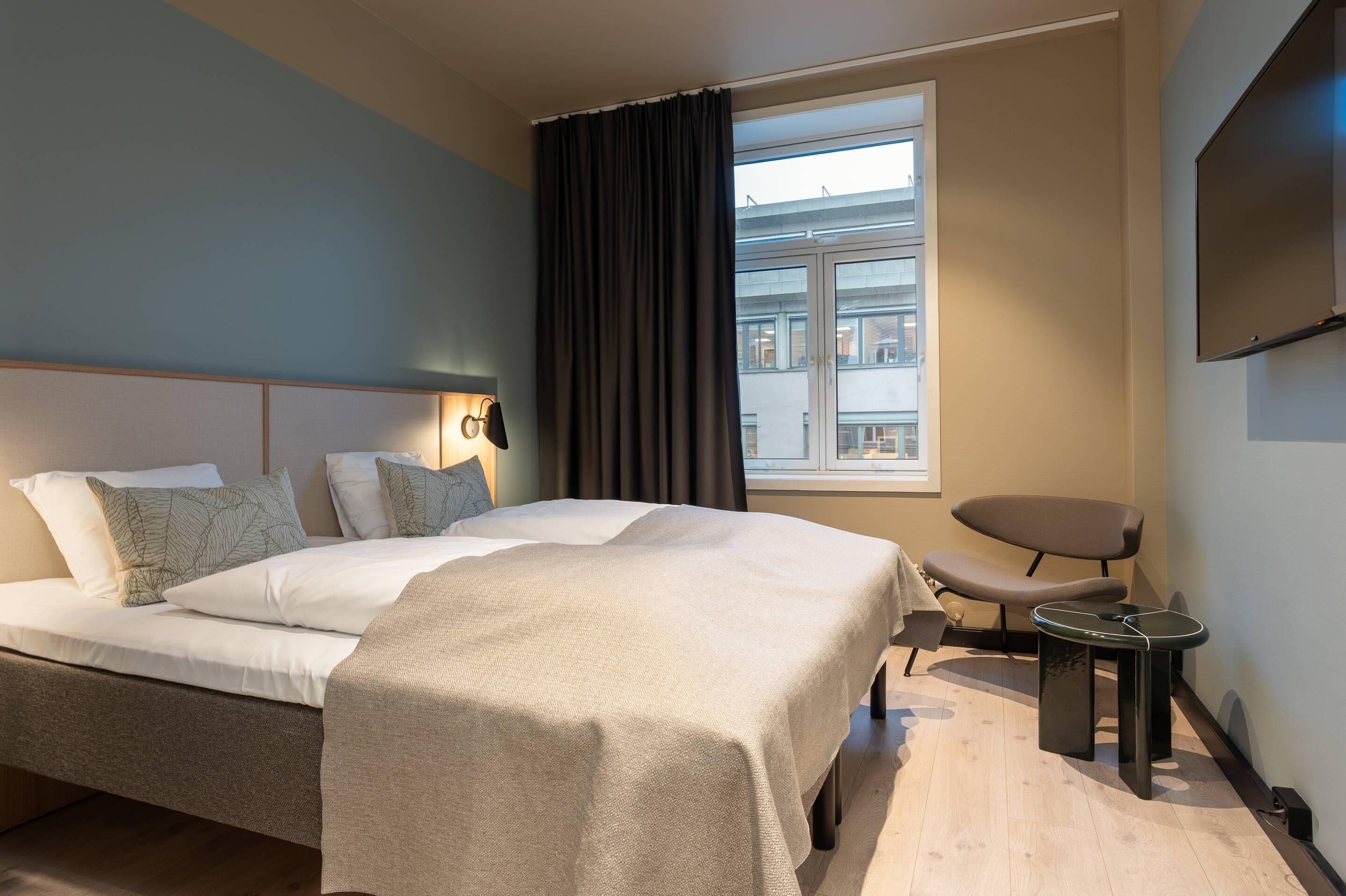 A hotel room with a bed, a chair, a small table, a TV, and a window with curtains.