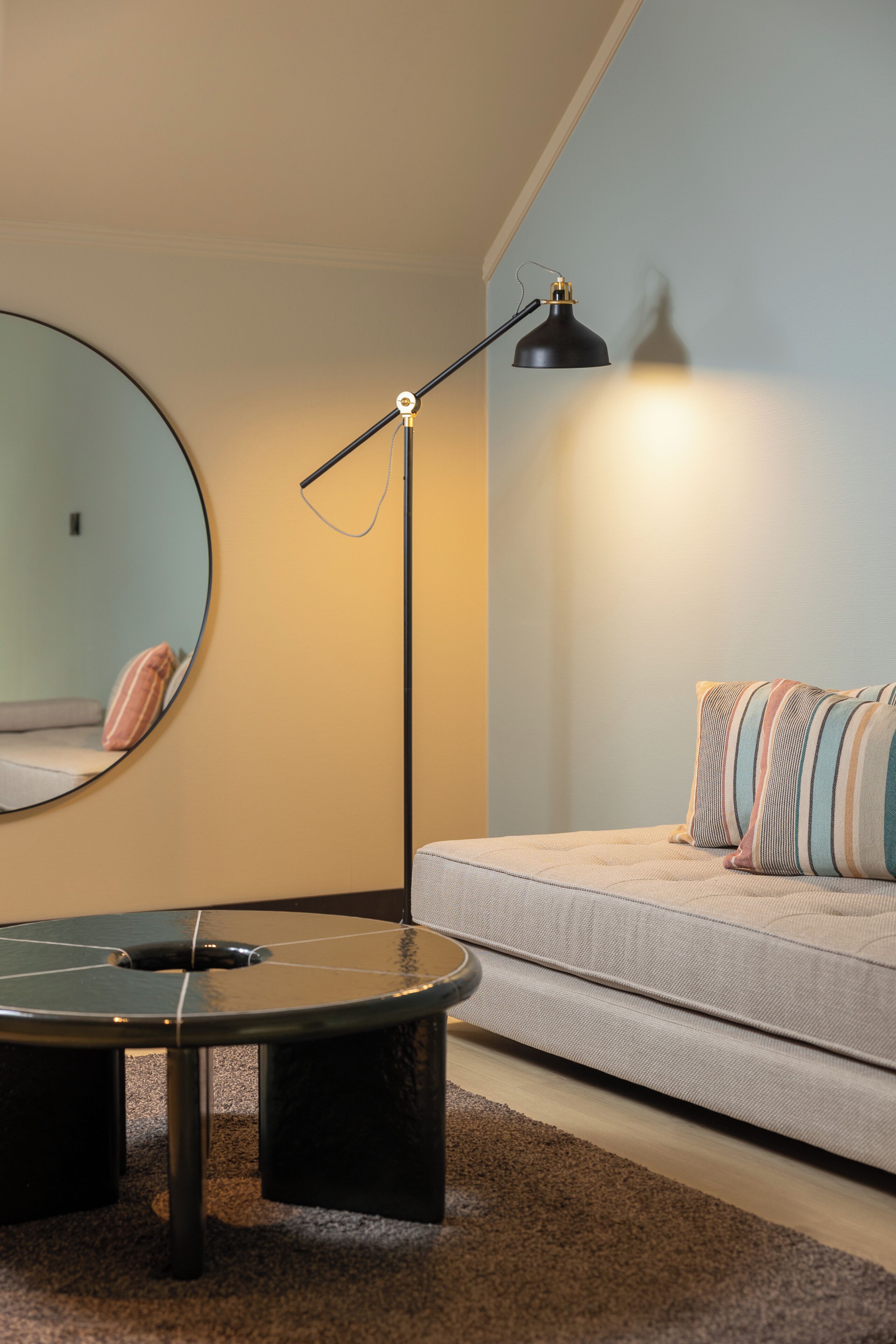 Een kamer met een ronde spiegel, een zwarte vloerlamp, een bank met gestreepte kussens, en een ronde salontafel.