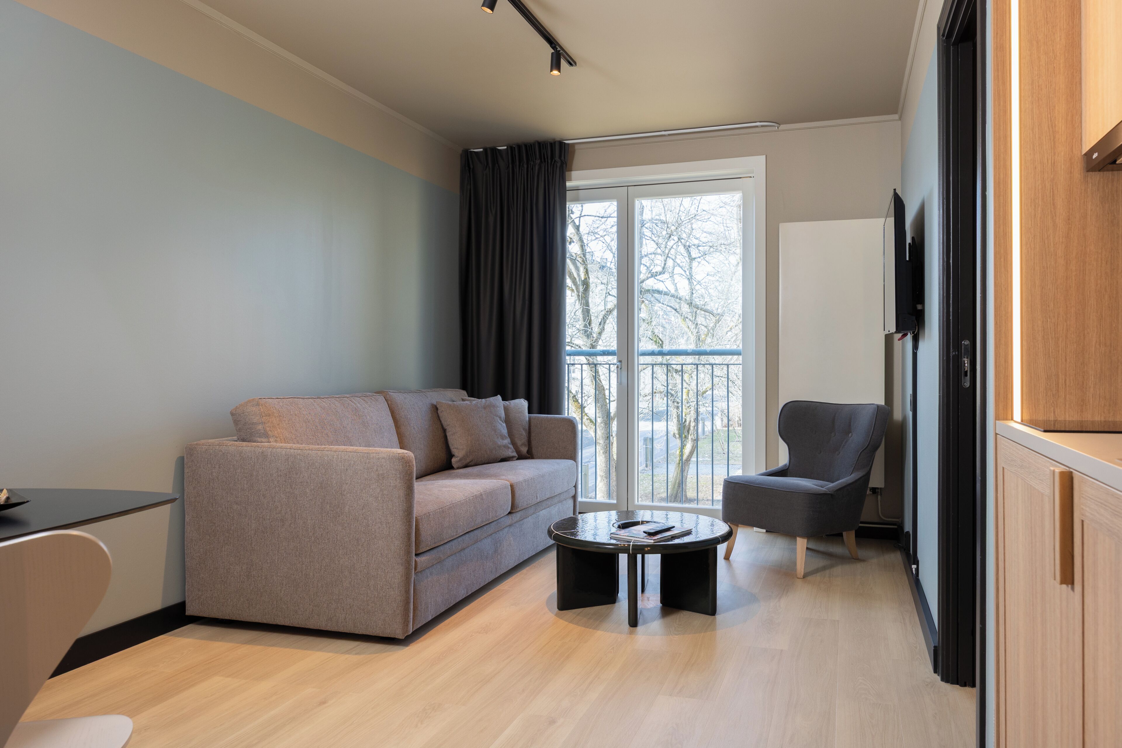 A modern living room with a sofa, armchair, and coffee table, a TV mounted on the wall, and a sliding glass door leading to a balcony.