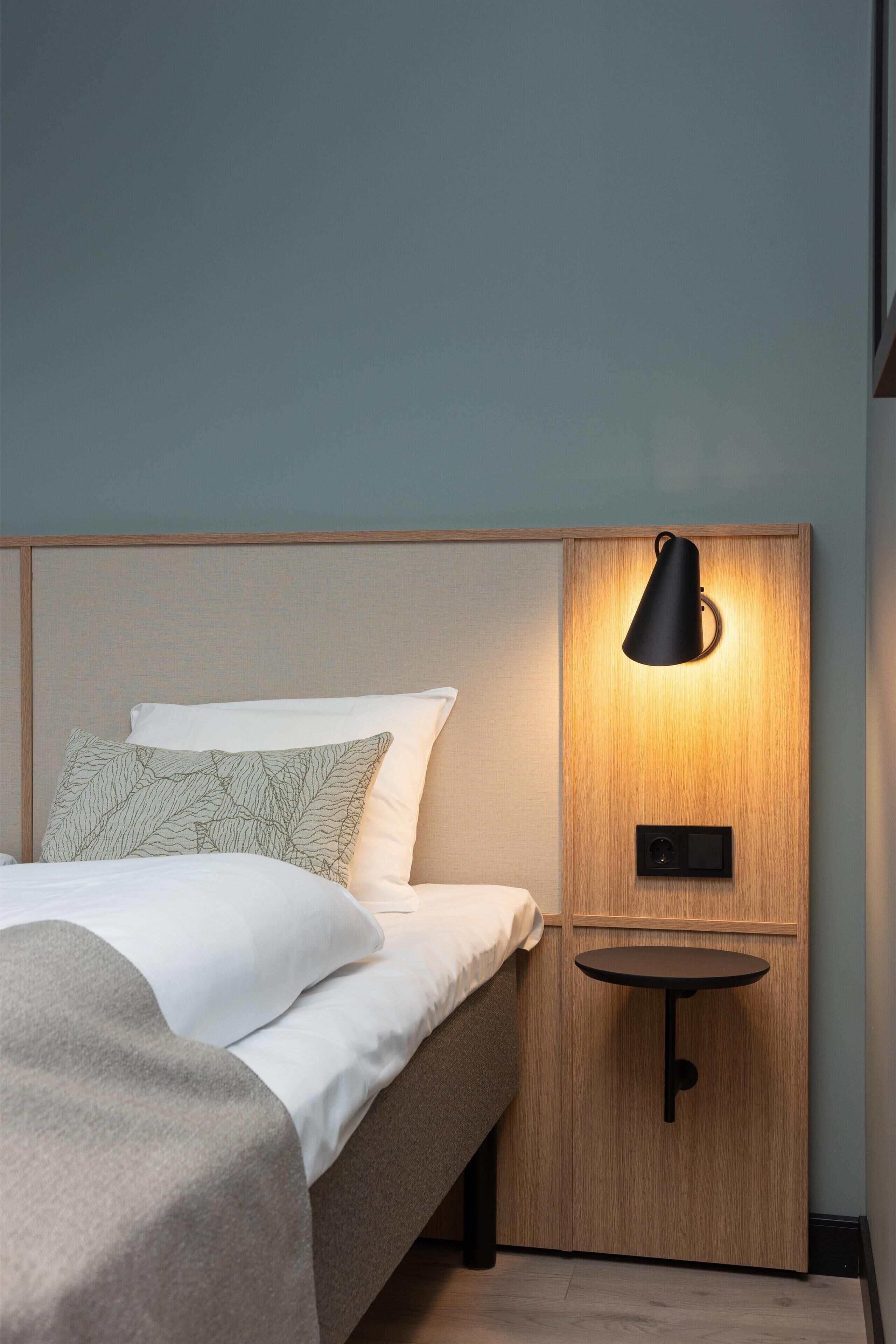 Ein ordentlich bezogenes Bett mit weißen Leinen und einer grauen Tagesdecke, ein Nachttisch mit einer schwarzen Lampe sowie eine Holzvertäfelung über dem Bett.