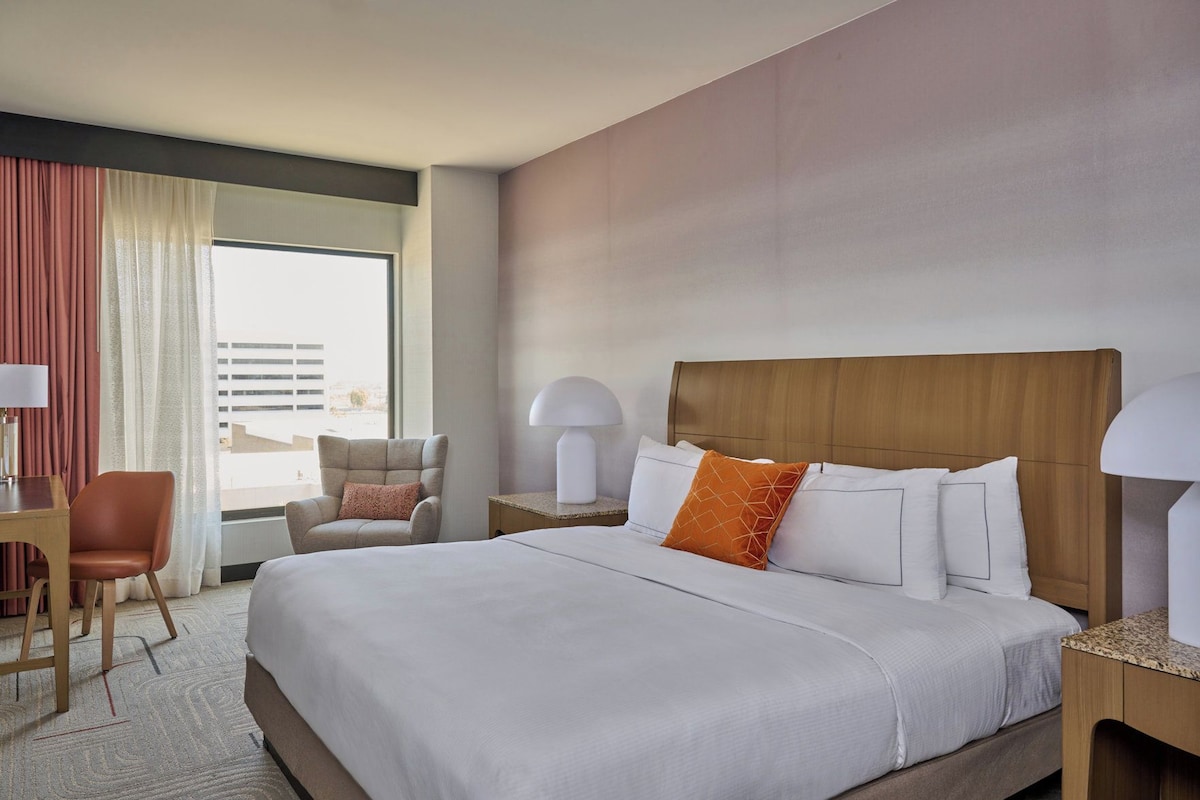 A hotel room with a large bed, a desk, a chair, and a window with curtains.