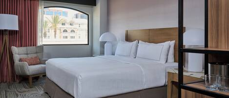 A hotel room with a large bed, a chair, a desk with a lamp, and a view of buildings through a window.