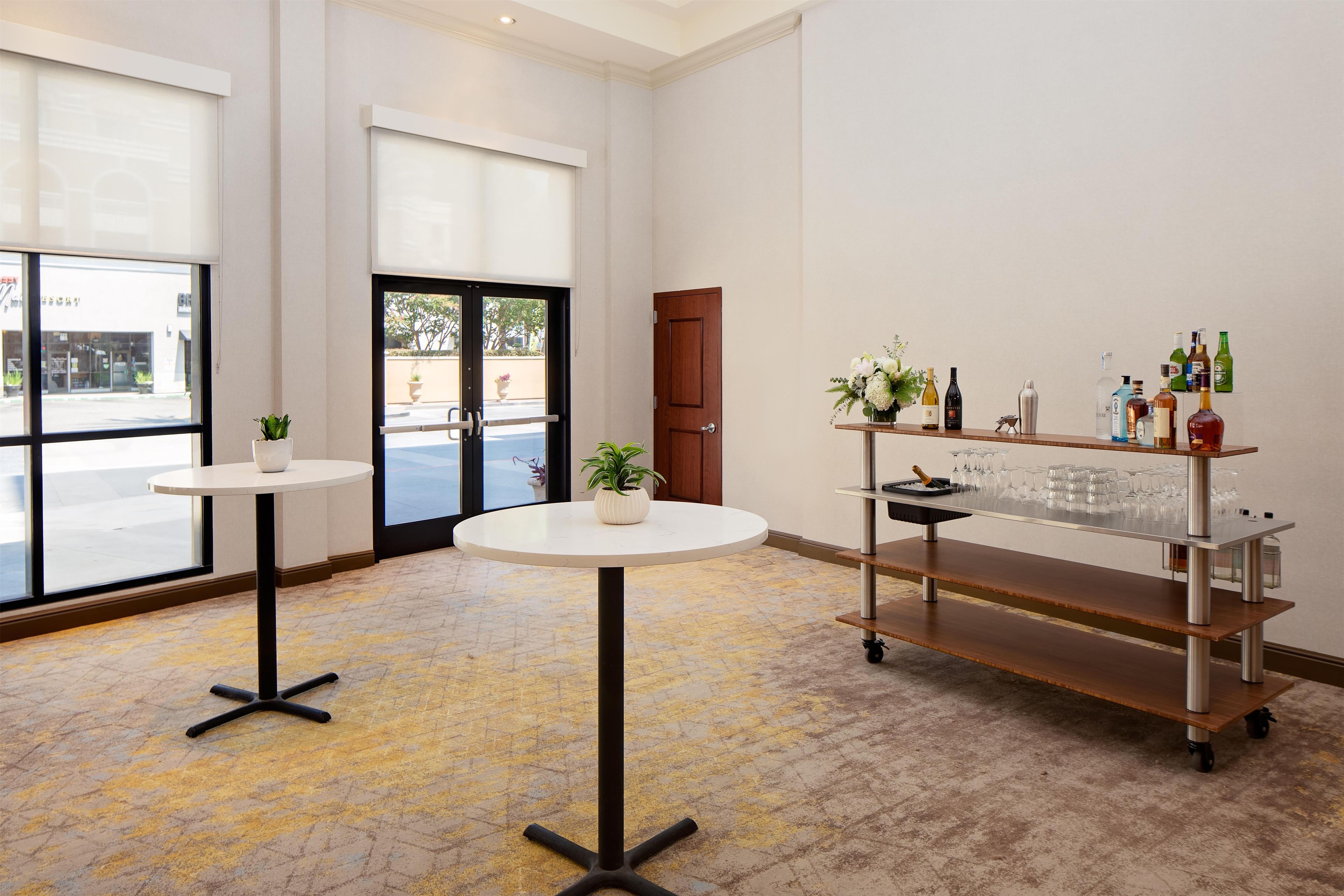 A hotel lobby with a round table, a potted plant, a bar cart with glasses, and a door leading outside.