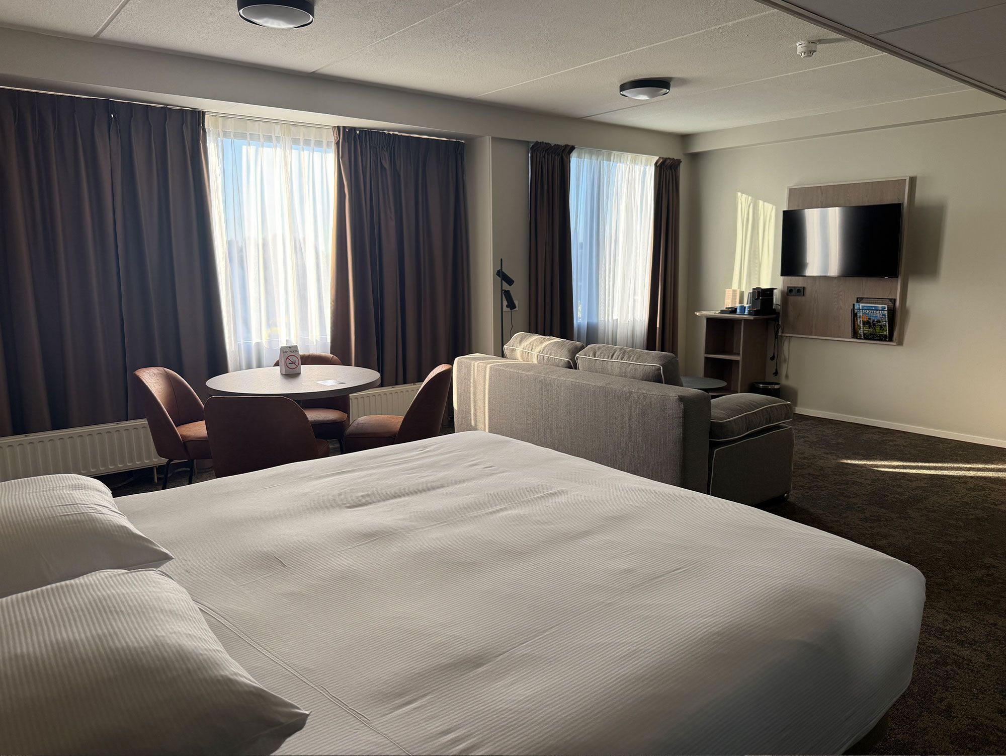 A hotel room with a large bed, a desk, a chair, a sofa, a TV, and a coffee machine.