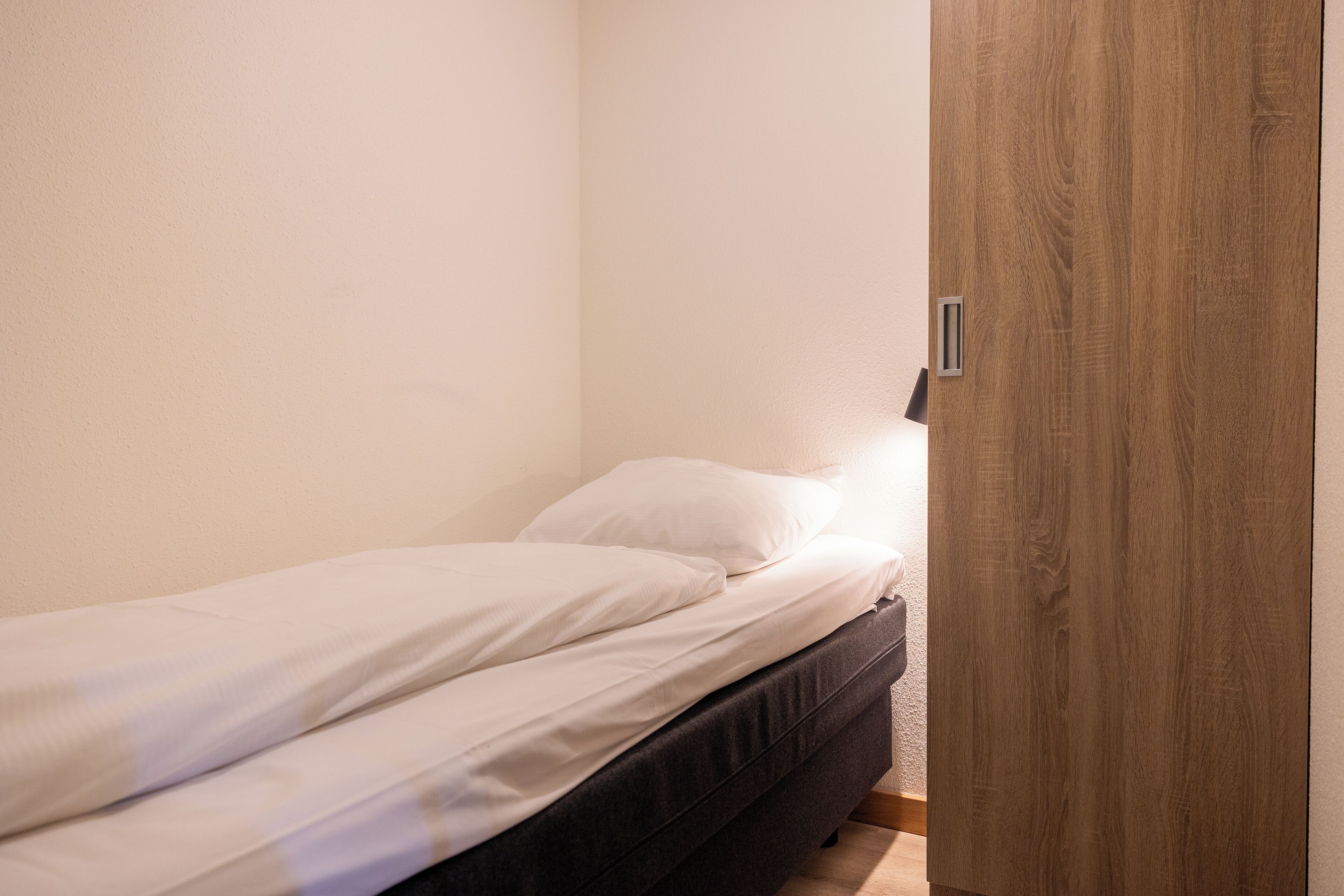 A small, neatly made bed with white linens in a corner room with a wooden door and a light switch on the wall.