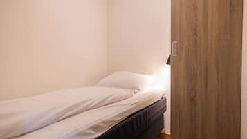 Un petit lit bien fait, avec du linge de lit blanc, dans une chambre d’angle dotée d’une porte en bois et d’un interrupteur sur le mur.