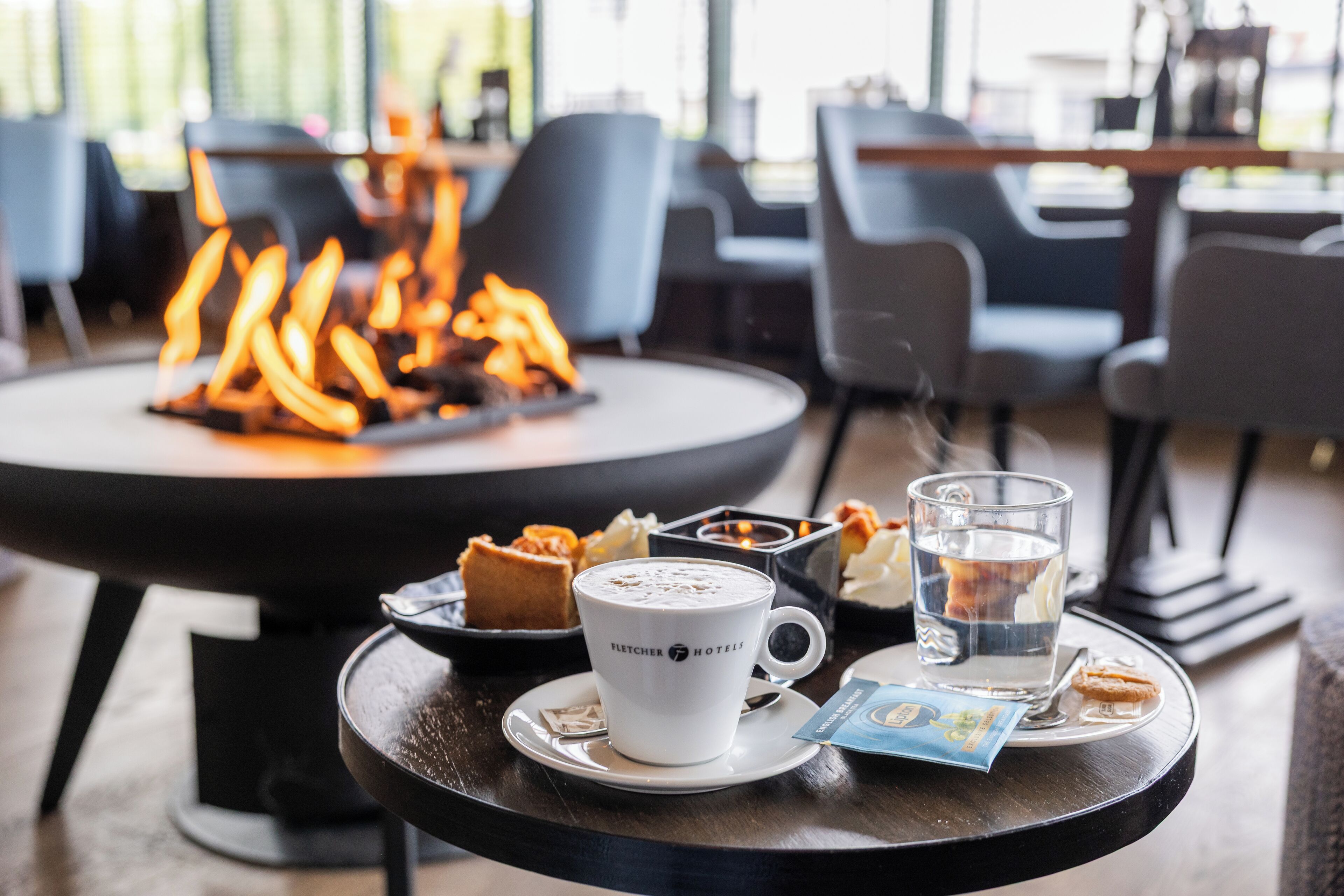 A table with a cup of coffee, a glass of water, and pastries, with a fire pit in the background.