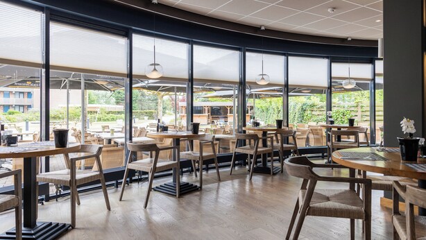 Een ruim café met grote ramen, houten tafels en rieten stoelen.