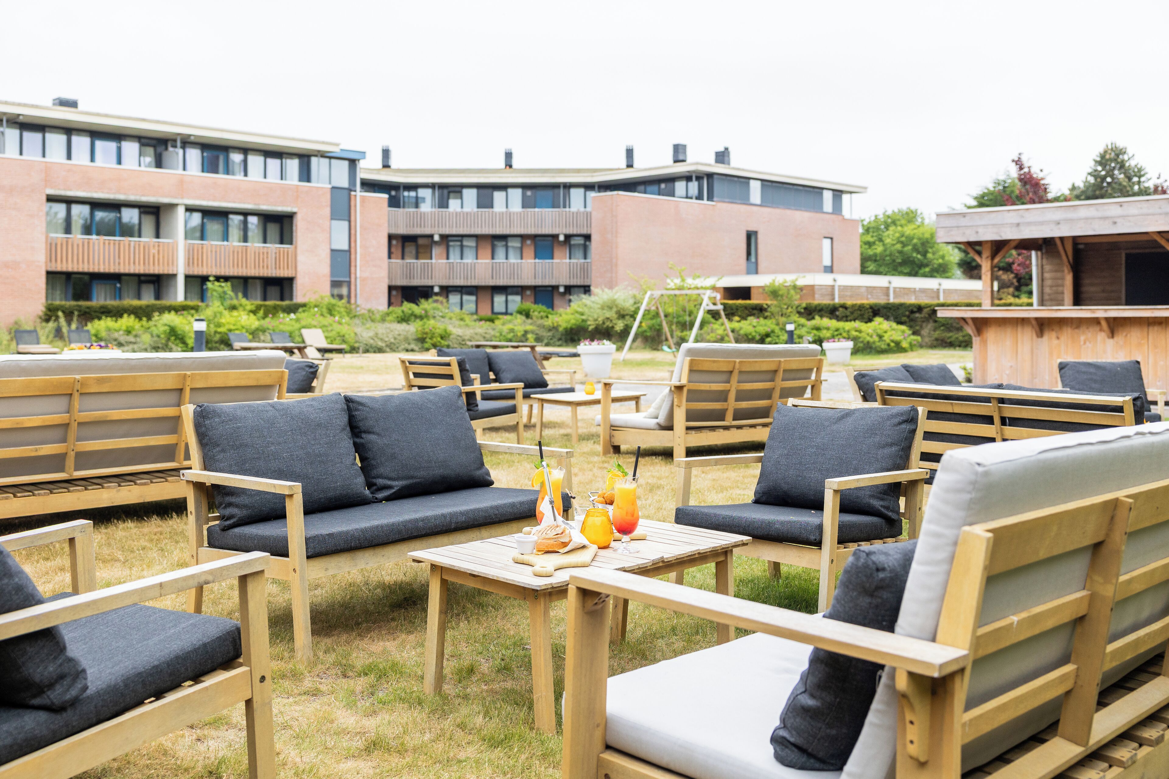 Un área de estar al aire libre con muebles y cojines de madera, una mesa con bebidas y aperitivos, y un edificio al fondo.