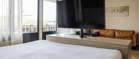 A hotel room with a large bed, a flat-screen TV mounted on the wall, a leather sofa, and a window with curtains.
