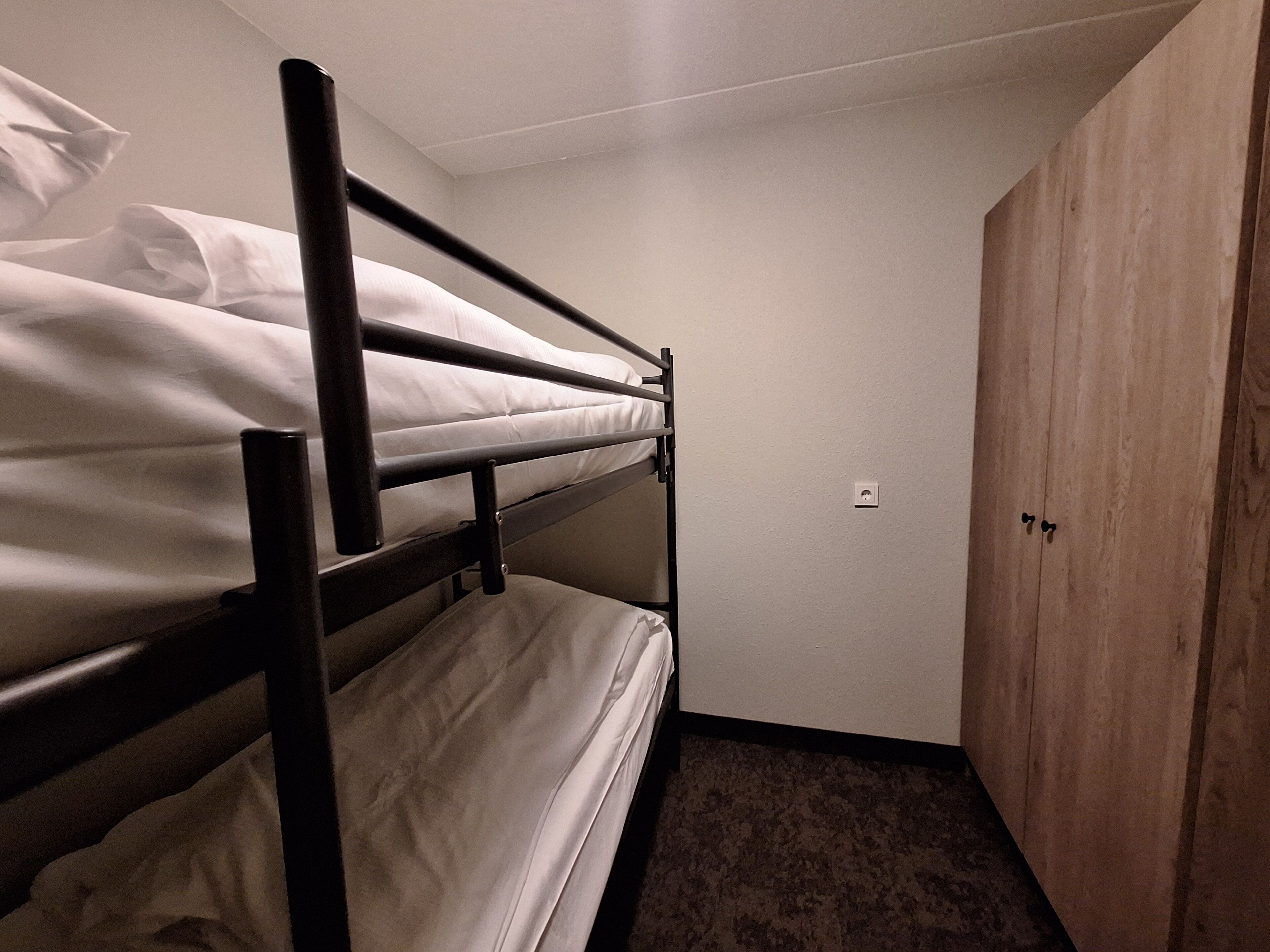 A bunk bed with white bedding, a wooden wardrobe, and a carpeted floor.