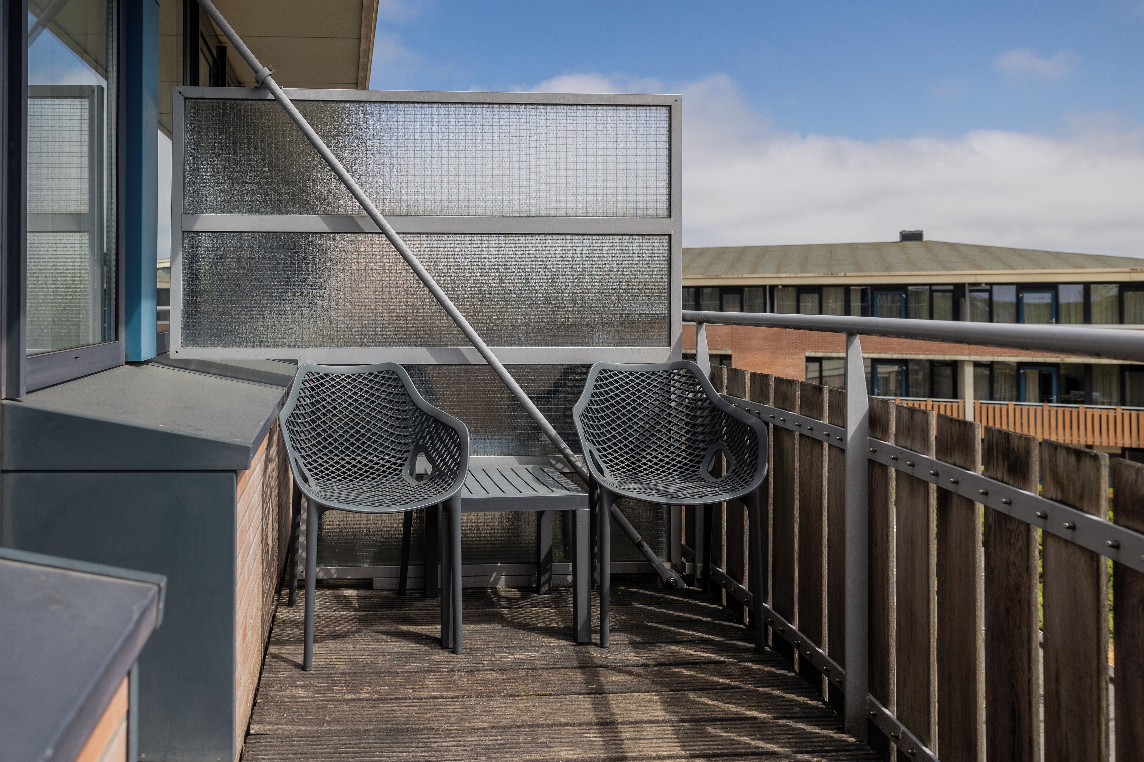 Un balcon avec une table et deux chaises en métal, une rambarde en métal et une vue sur un immeuble doté d’un toit végétalisé.