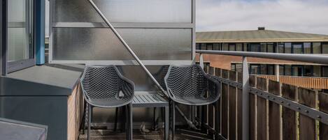 Un balcon avec une table et deux chaises en métal, une rambarde en métal et une vue sur un immeuble doté d’un toit végétalisé.