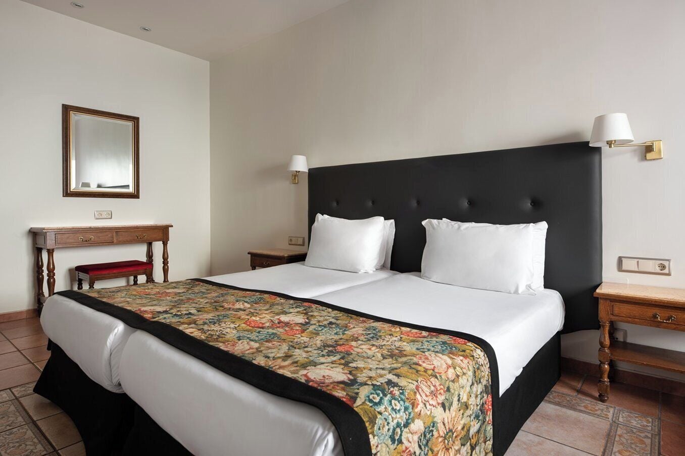 A hotel room with a large bed, two bedside tables, a nightstand, a wooden bench, a desk, and a mirror.