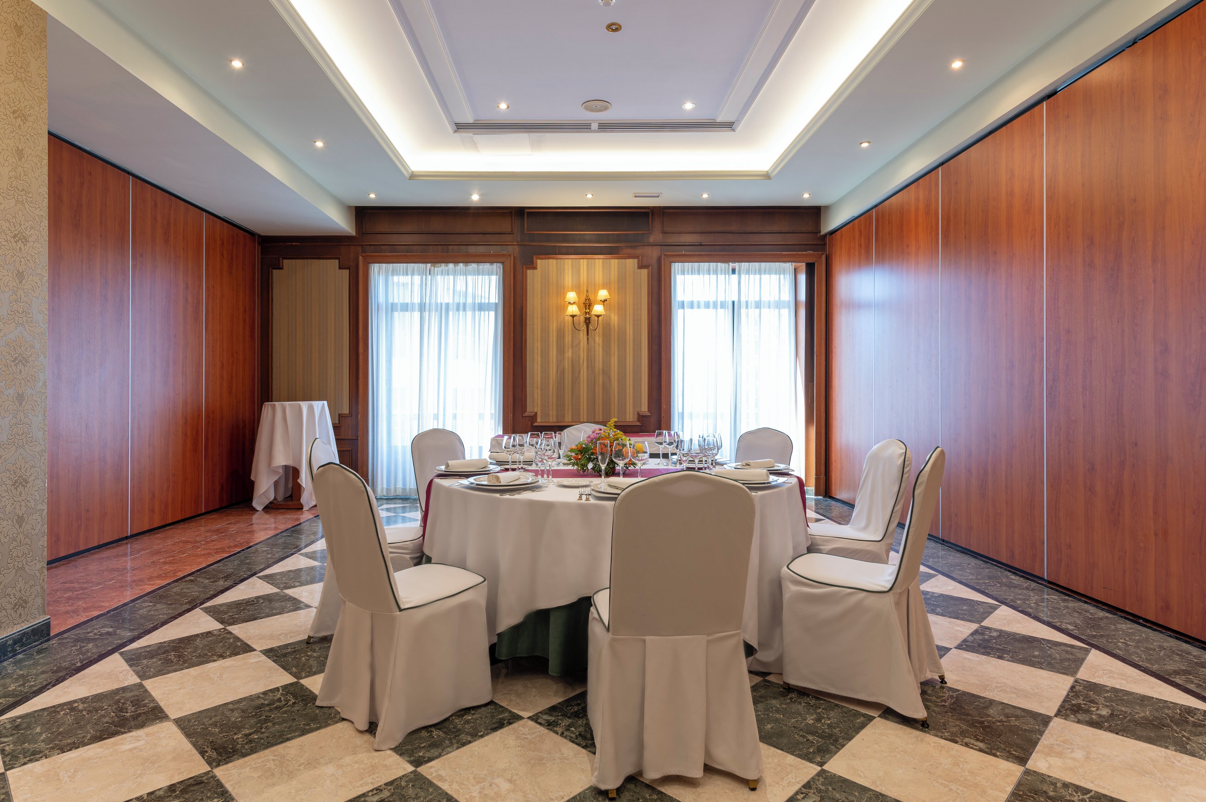 A conference room with a round table set for a meeting, surrounded by chairs, with a checkered floor and wooden panel walls.