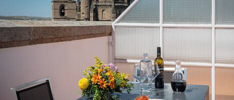 A table with a plate of food, a bottle of wine, and a vase of flowers on a balcony with a view of a historic building.
