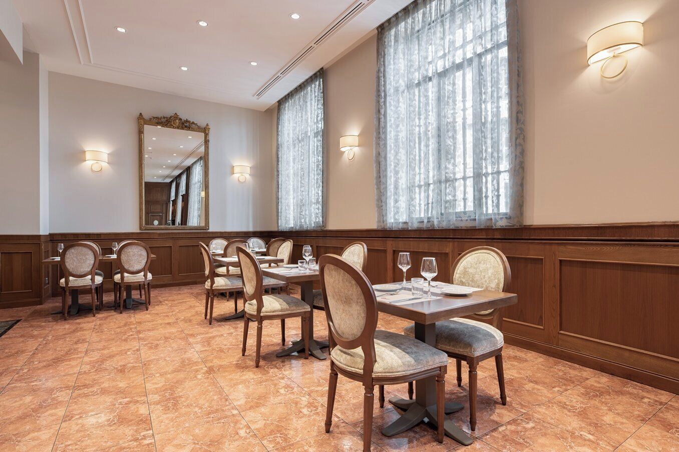 Un comedor de hotel con mesas y sillas de madera, ventanas amplias con cortinas translúcidas y lámparas fijadas a la pared.