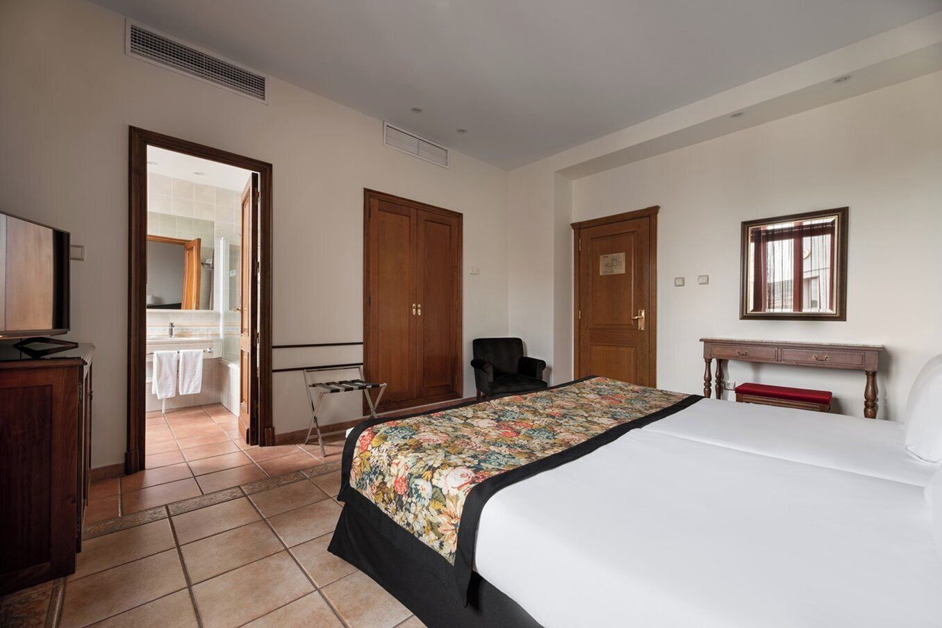 Habitación de hotel con cama, escritorio, silla, armario y baño visible a través de una puerta abierta.
