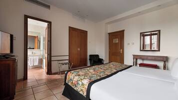Habitación de hotel con cama, escritorio, silla, armario y baño visible a través de una puerta abierta.