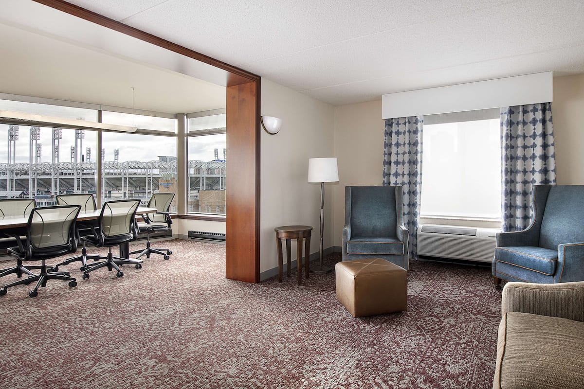 A hotel room with a conference table, office chairs, a sofa, a small ottoman, a lamp, and a large window with curtains.