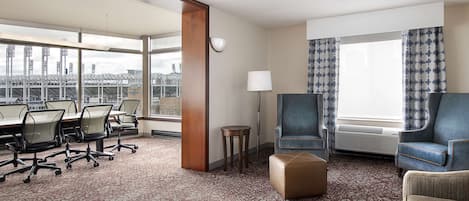 Quarto de hotel com uma mesa de conferência, cadeiras de escritório, um sofá, um pequeno banquinho, uma luminária e uma grande janela com cortinas.