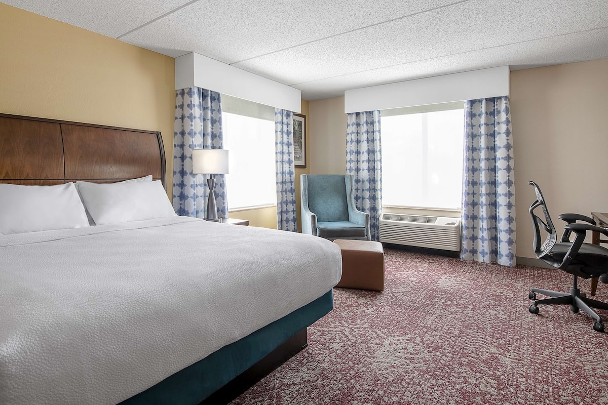 A hotel room with a large bed, a desk with a chair, a window with curtains, and a wall-mounted air conditioning unit.