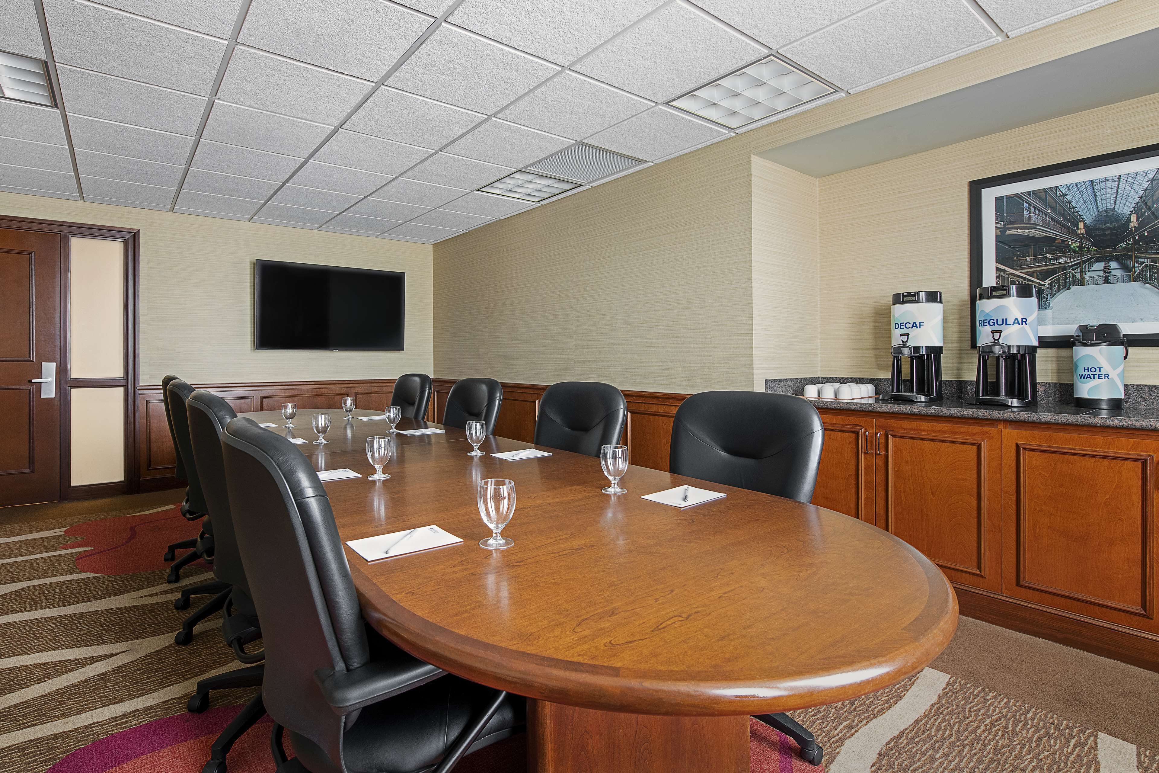 A conference room with a wooden table, black chairs, a flat-screen TV on the wall, and a coffee station with decaf, regular, and hot water options.