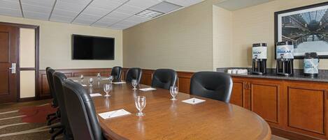 A conference room with a wooden table, black chairs, a flat-screen TV on the wall, and a coffee station with decaf, regular, and hot water options.