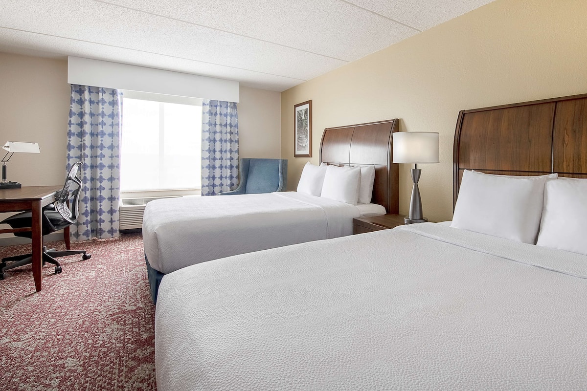 A hotel room with two beds, a desk, a chair, a lamp, and a window with curtains.