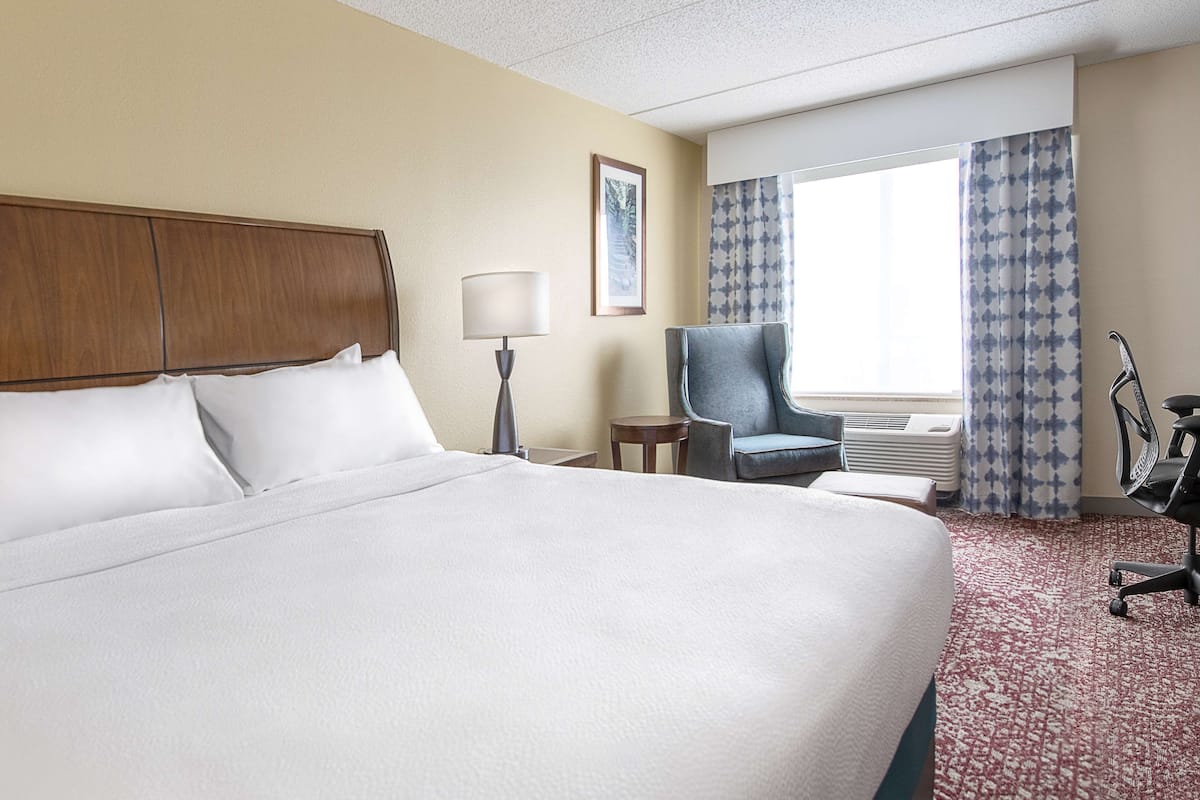A hotel room with a large bed, a chair, a desk, and a window with curtains.
