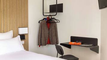 A hotel room with a bed, a chair, a desk with a laptop, a TV mounted on the wall, and a coat rack with hangers.