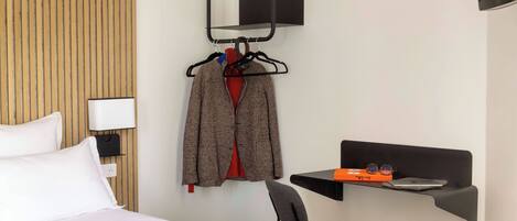 A hotel room with a bed, a chair, a desk with a laptop, a TV mounted on the wall, and a coat rack with hangers.