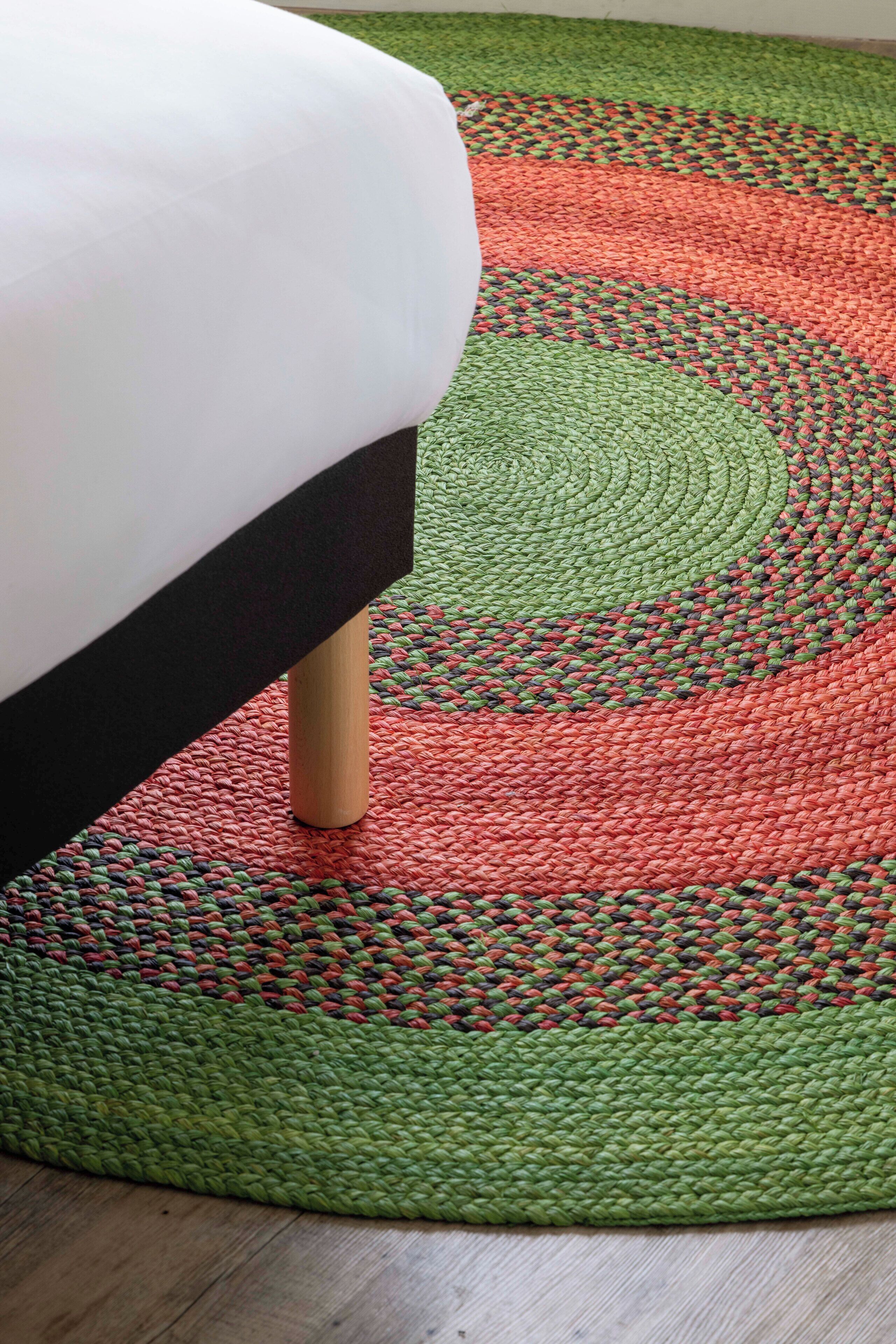 A bed with a white headboard and wooden legs, positioned on a green and red woven rug with circular patterns.