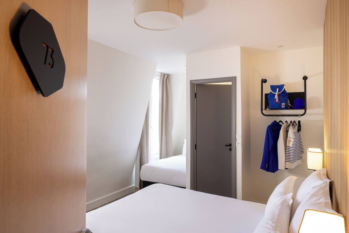 A hotel room with a bed, a door, a coat rack with clothes, and a wall clock.