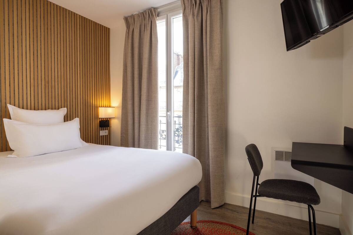 A hotel room with a large bed, a desk, a chair, a TV, and a window with curtains.