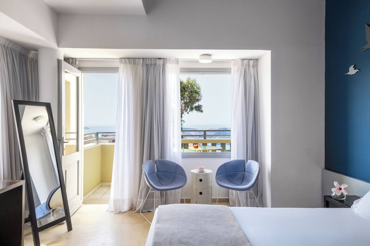 Une chambre d’hôtel avec un lit, deux chaises, une petite table et un balcon donnant sur la mer.