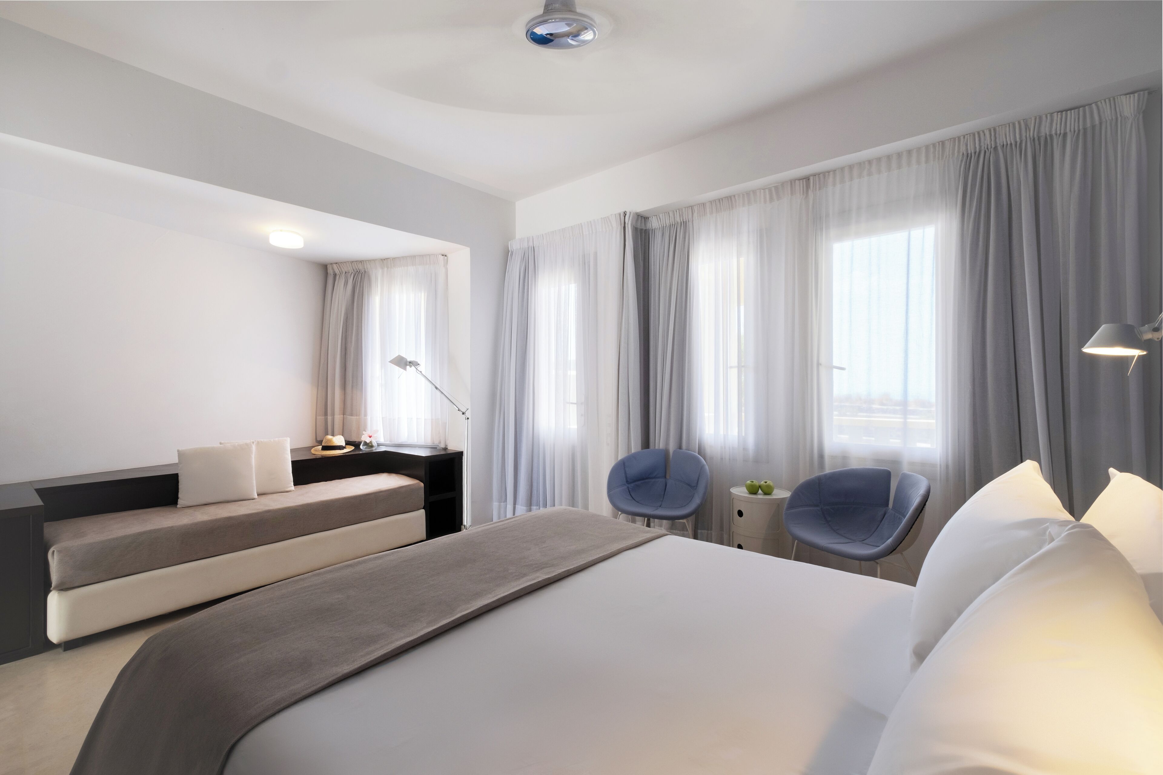 A modern hotel room with a large bed, two armchairs, a desk with a lamp, and a view of the city through large windows.