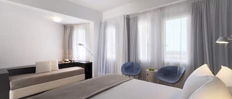 Een moderne hotelkamer met een groot bed, twee fauteuils, een bureau met lamp en uitzicht op de stad door grote ramen.