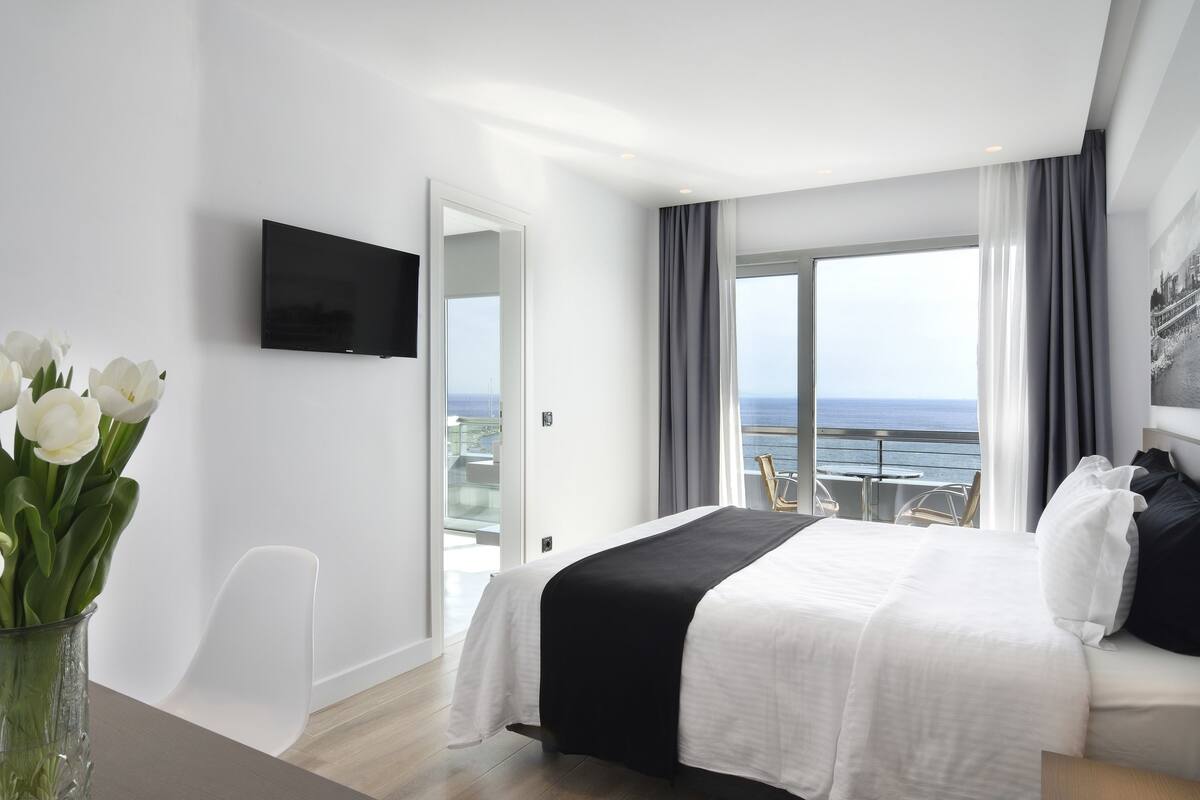 A modern hotel room with a large bed, a flat-screen TV, a vase of white flowers, and a balcony with a view of the sea.