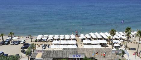 A beachside restaurant with outdoor seating and umbrellas, a parking area, and a view of the sea.